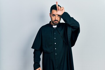 Handsome hispanic man with beard wearing catholic priest robe making fun of people with fingers on...