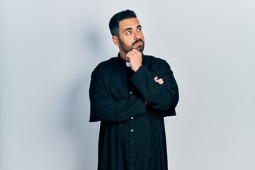 Handsome hispanic man with beard wearing catholic priest robe with hand on chin thinking about question, pensive expression. smiling with thoughtful face. doubt concept.