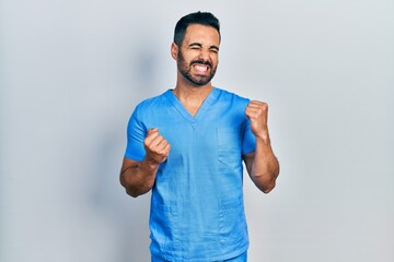 Handsome hispanic man with beard wearing blue male nurse uniform excited for success with arms raised and eyes closed celebrating victory smiling. winner concept.