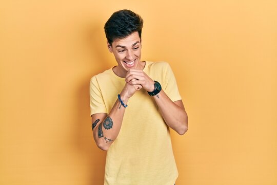 Young hispanic man wearing casual yellow t shirt laughing nervous and excited with hands on chin looking to the side