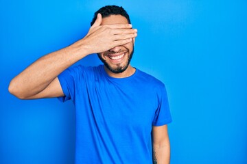 Hispanic man with beard wearing casual blue t shirt smiling and laughing with hand on face covering eyes for surprise. blind concept.
