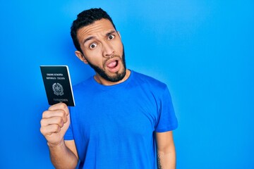 Hispanic man with beard holding italy passport in shock face, looking skeptical and sarcastic, surprised with open mouth