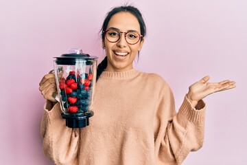 Young hispanic girl holding food processor mixer machine with fruits celebrating achievement with...