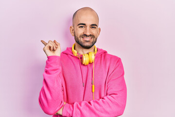 Obraz premium Young bald man wearing gym clothes and using headphones with a big smile on face, pointing with hand and finger to the side looking at the camera.