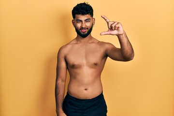 Fototapeta premium Arab man with beard wearing swimwear shirtless smiling and confident gesturing with hand doing small size sign with fingers looking and the camera. measure concept.
