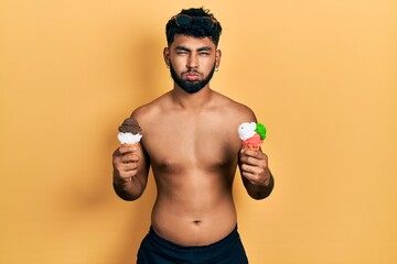Fototapeta premium Arab man with beard wearing swimwear eating two ice cream cones puffing cheeks with funny face. mouth inflated with air, catching air.