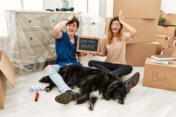 Young caucasian couple with dog holding our first home blackboard at new house surprised with hand...