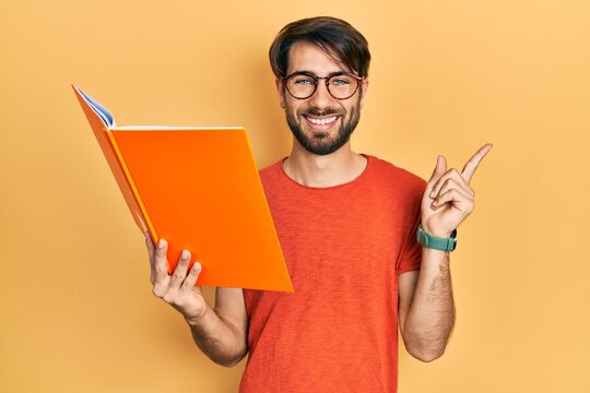 Young hispanic man reading book smiling happy pointing with hand and finger to the side