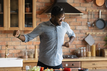 Happy excited Black hipster guy having fun in kitchen, cooking salad, dancing at table with sliced fresh vegetables, enjoying household activities, preparing lunch, dinner, eating healthy organic food