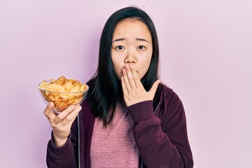 Young chinese girl holding potato chip covering mouth with hand, shocked and afraid for mistake. surprised expression