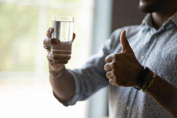 Satisfied mixed raced Black young man approving drinking fresh water quality, holding transparent glass, making like hand gesture, showing thumb up at camera. Close up of hands, cropped shot