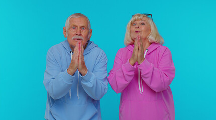 Please, God, forgive us. Senior old grandfather grandmother sincerely praying to God and looking up with pleading expression, begging apology, beseeching with hopeful face on blue studio background