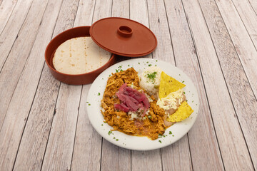 Plate of cochinita pibil stew with white rice, grated cheese, stewed pull chicken meat, corn chips, chopped fresh parsley and hot wheat tortillas on wooden table