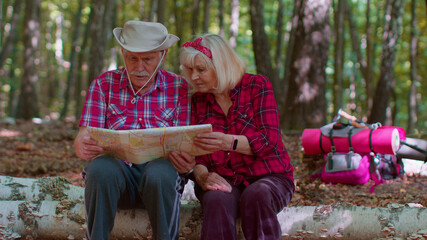 Old married couple sitting in forest and examining route plan. Senior man woman lost in wood, searching way back. Tourists looking at map while hiking. Active mature family leisure. Tourism traveling