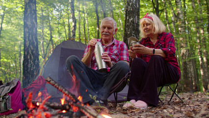 Senior elderly grandmother grandfather drinking tea over campfire in wood. Old man woman tourists having rest pouring hot drink from thermos at camping in forest. Active rest outdoors of mature family