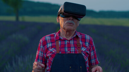 Senior grandfather farmer in virtual reality helmet in field of lavender flowers. Elderly man wearing VR glasses for digital management of greenhouse cultivation. VR technologies modern agribusiness