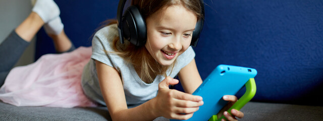 Banner of Cute little girl in headphones lying on couch with on digital tablet computer, Kid addicted to technology