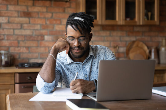 Focused Black mixed race student writing notes, watching and listening webinar on laptop, summarizing online lecture, attending virtual class. Remote worker in glasses working at home office workplace