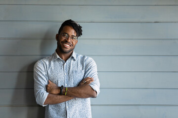 Head shot of handsome young African American guy, young Black man with dreads wearing casual stylish shirt, glasses, keeping hands folded, smiling at camera. Male portrait with copy space