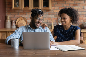 Happy African American couple paying monthly bills, insurance, mortgage fees online on laptop, using computer at home, shopping on internet, getting and discussing good news from online chat - Powered by Adobe