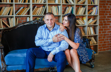 stylish man in a blue shirt with a beautiful wife in a dress are sitting together on a cozy sofa against the background of a luxurious room with books. Family happiness