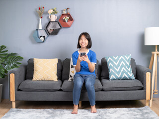 asian girl sit on couch in living room holding smart phone, chatting use social media network, watch received video from friend, with modern wireless technology.