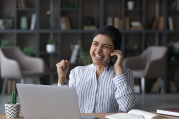 Excited young Indian woman sit at desk at home office work on laptop get promotion offer on...