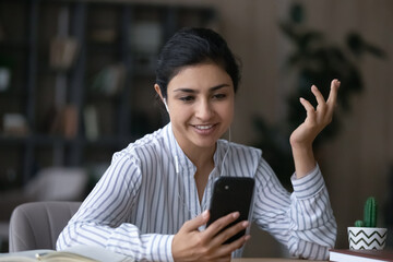 Smiling young Indian woman in headphones talk speak on video call on cellphone gadget. Happy mixed race female in earphones have webcam virtual digital event on smartphone. Communication concept.