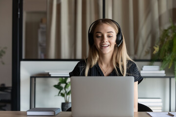 Smiling young Caucasian female employee worker use laptop wear headphones have webcam digital virtual event. Happy millennial woman coach in earphones talk speak on video call on computer in office.