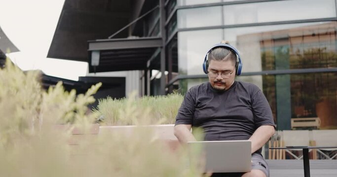 Obese Asian man with headphone sit on using laptop working outdoors in garden. Meeting video call with colleagues. Freelance online stock trading. Work online with free internet signal. 