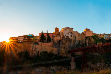 Naklejka premium Cityscape in old town changing light at sunset; Hanging houses with old town hanging over mountain and red bridge of San Pablo de Cuenca with sun star, world heritage site, Spain. Horizontal view