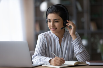 Millennial Indian female student in headphones handwrite make notes study online on webcam video call on computer at home. Smiling young mixed race woman in earphones write work distant on laptop.
