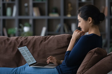 Happy millennial Indian woman sit relax on couch use computer study online from home. Smiling young...