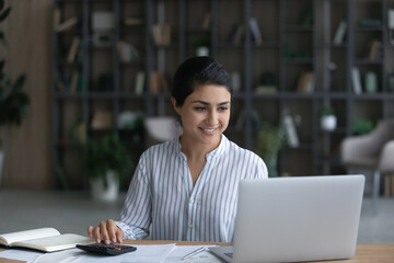 Happy young Indian woman sit at desk pay bills online on laptop, calculate expenses expenditures....