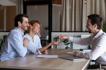 Smiling male realtor or broker give keys to exited Caucasian couple buyers close deal make agreement at meeting. Happy agent congratulate greet smiling man and woman buy first home together.