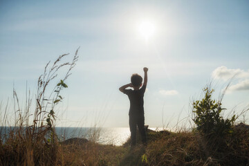 Healthy fit boy raising his hand after reaching to mountain summit by hikking. Sporty young child looking at sea and sunset on top of hill cliff. Success goal for outdoor sport activity.