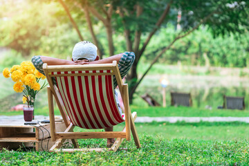 Back view of senior man with white hat sitting on garden chair and by the table in garden. Summer vacation in green surroundings. Happy person outdoors relaxing on deck chair in garden.Outdoor leisure
