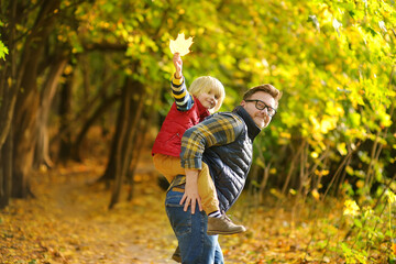 Little boy and his father having fun during stroll in the forest on sunny autumn day. The father rides the child on own back. Hiking with kids.