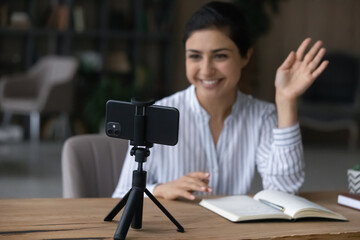 Smiling young Indian woman wave greet talk speak on video call on cellphone at home office. Happy mixed race ethnicity female coach or trainer have webcam lesson or virtual event on smartphone.