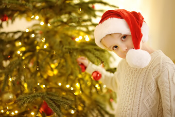 Little boy wearing Santa hat ready for celebrate Christmas. Cute child decorating the Christmas tree with glass toy. Baby hopes of magic and gifts at New Year night.