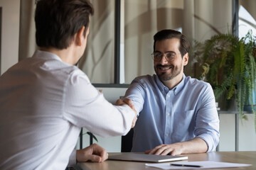 Smiling young Caucasian businessman handshake male colleague or job candidate at meeting in office....