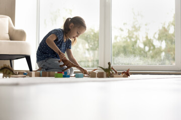 Focused little kid engaged in game with toy dinosaurs, wooden construction blocks. Preschooler girl playing on heating warm floor at window. Safe house, child skills development, childhood