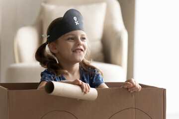 Happy excited little kid in playing pirate at home. Funny child wearing black sailor hat with skull sailing in cardboard box boat, enjoying adventure, having fun. Childhood, fantasy, costume party
