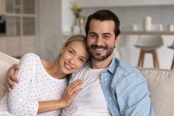 Happy millennial married couple of homeowners head shot portrait. Husband and wife sitting on couch, looking at camera with toothy smile, hugging with love, relaxing in new comfortable house