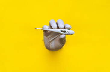 A hand in a white medical glove is pushed through a torn hole in yellow paper and holds an electronic thermometer. The concept of diagnosing colds, covid-19, coronavirus. Copy space.