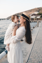 Happy cheerful baby girl sitting on mother hands wear stylish white dresse and straw hat relax over sea shore at quayside outdoors close up. Looking at camera. Motherhood. Maternity.