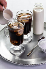 hand pouring cream into an ice coffee drink with a bottle of milk in behind on a metal tray.