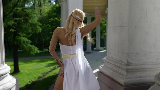 a blonde in a Greek chiton is dancing among the columns. view from the back. the camera moves away