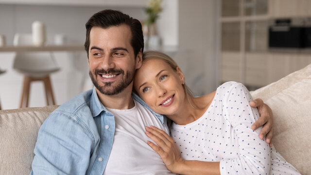 Smiling millennial married couple relaxing and hugging on couch at home, looking away with happy faces, planning mortgage, dreaming of own house, thinking of future vision together. Banner image