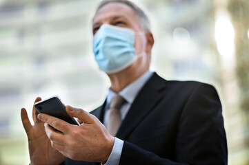 Senior manager using his smartphone outdoor while wearing a mask to protect from Coronavirus pandemic
